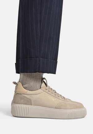 Beige snøresneakers med tyk sål båret med lysebeige sokker og navy blå, pinstripe bukser med opbuk på en hvid baggrund.