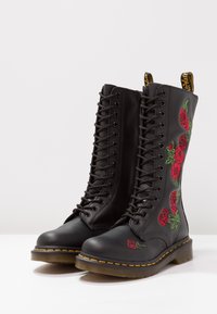 Bottes noires en cuir avec lacets, ornées d'une broderie de roses rouges, avec un col rembourré et des coutures jaunes sur la semelle. Texture lisse et design haut.