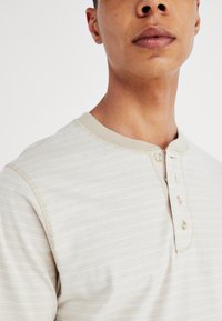 Svetlo béžová henley košeľa s jemným prúžkovaným vzorom, krátkymi rukávmi a gombíkovou légou. Mäkká textúra s rebrovaným golierom.