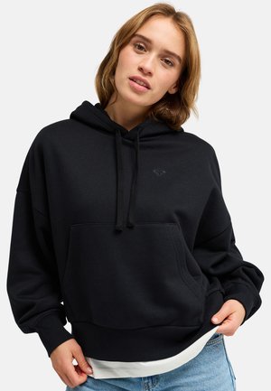 Jeune femme aux cheveux châtain clair portant un sweat à capuche noir et un jean bleu, tenant l'ourlet du sweat d'une main.