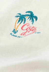 Motif brodé sur tissu blanc avec deux palmiers et le texte « Côte d'Azur Plage de Ville d'Ail » en fil bleu et rose.