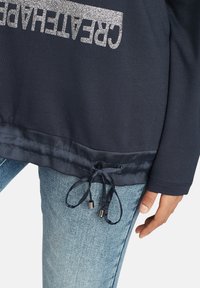 Marinblå sweatshirt med metallisk silvertext "KREATIVITET", med satingrim och dragsko med silverfärgade spetsar, kombinerad med ljusblå jeans.