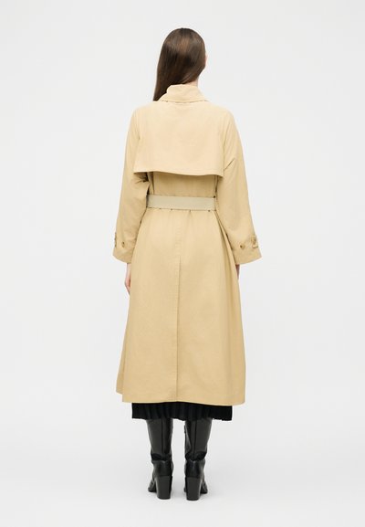 Femme aux longs cheveux bruns portant un trench-coat beige ceinturé, une jupe noire et des bottes à talons noires, debout de dos sur un fond uni.
