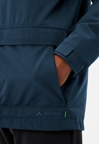 Giacca impermeabile blu marina con una texture liscia, polsini regolabili e chiusura in Velcro, con un logo e un accento verde sull'orlo.
