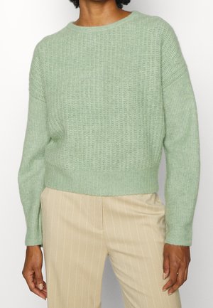 Person trägt einen hellgrünen, strukturierten Strickpullover mit langen Ärmeln und beige-gestreifte Hosen vor einem einfarbigen Hintergrund.