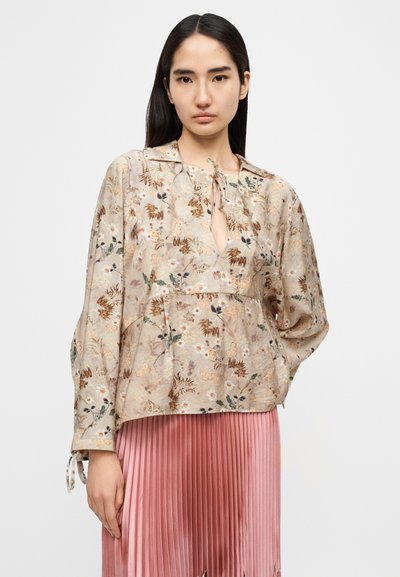 Μπεζ μπλούζα με λουλουδάτο μοτίβο και μακριά μανίκια, με λεπτομέρεια δέσιμου στη λαιμόκοψη, και ροζ φούστα με πιέτες, που παρουσιάζει ένα στρωματοποιημένο, υφασμάτινο στυλ.