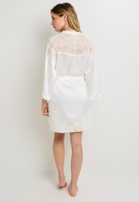 Accappatoio bianco di satin con dettagli in pizzo sulle spalle e sull'orlo, dotato di una vita cinturata e di una finitura liscia e lucida.