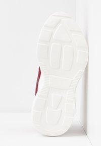 Semelle d'une chaussure en caoutchouc blanche avec des motifs de bande de roulement appuyée contre un mur d'angle blanc, montrant une partie de la partie supérieure rouge.