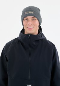 Donkerblauwe waterdichte jas met een hoge kraag, een rits aan de voorkant en ventilerende delen, gecombineerd met een grijze beanie met een logo.