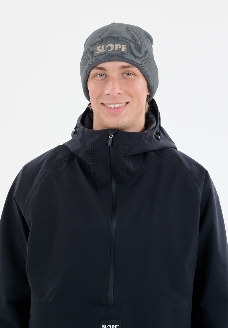 Donkerblauwe waterdichte jas met een hoge kraag, een rits aan de voorkant en ventilerende delen, gecombineerd met een grijze beanie met een logo.