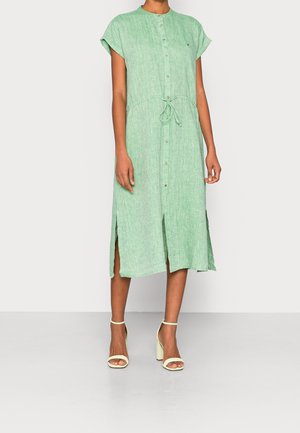 Kvinde iført en lysegrøn midi-kjole med knapper foran, korte ærmer, bindebånd i taljen, sideslids og cremefarvede sandaler med blokhæl.