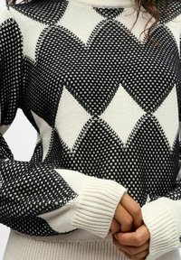 Sweter dzianinowy z czarno-białym wzorem w diamenty, z prążkowanymi mankietami i teksturowanym wykończeniem. Materiał wydaje się miękki i ciepły.