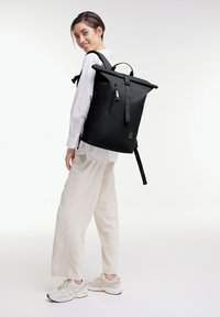 Mustan rullattavan repun, joka on valmistettu kestävästä materiaalista. Repussa on säädettävät hihnat, etutasku vetoketjulla ja minimaalinen logokoristus.