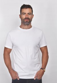 T-shirt bianca a maniche corte con scollo rotondo, realizzata in tessuto di cotone lucido. Il modello indossa jeans scuri e un bracciale nero.