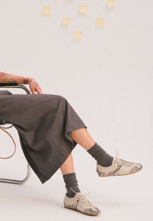 Henkilö istuu tuolissa, yllään harmaa hame, tummanharmaat sukat ja beigeä käärmeen nahkakuviolla koristellut nauhalliset kengät. Kädessä näkyy tatuointeja.