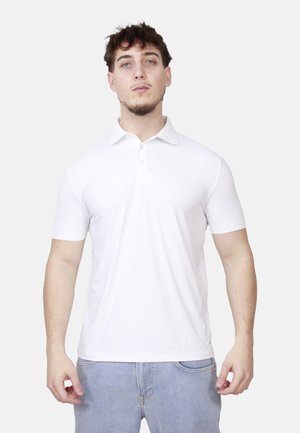 Giovane uomo con capelli corti e ricci che indossa una polo bianca a maniche corte e jeans azzurri, in piedi davanti a uno sfondo neutro.