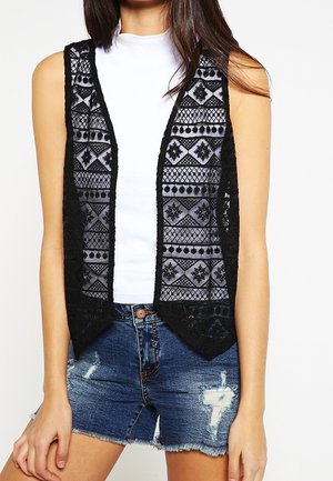 Veste sans manches en dentelle noire avec des motifs géométriques sur un top blanc sans manches, associée à un short en denim bleu délavé.