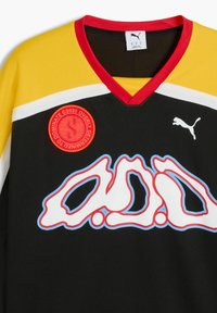 Čierny športový dres s žltými rukávmi, červenými akcentmi a bielymi pruhmi. Obsahuje veľký grafický dizajn v bielej a modrej farbe s kruhovým logom.