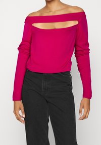 Pull magenta à épaules dénudées avec manches longues, présentant un design découpé sur la poitrine et un tissu lisse et léger.