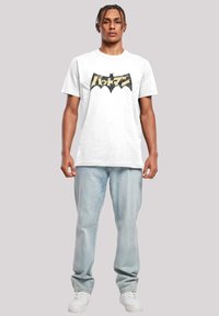 Hvit bomulls t-skjorte med et svart og beige flaggermus-symbolgrafikk, kombinert med lyseblå jeans og hvite sneakers.
