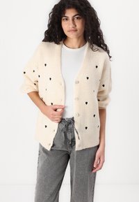 Kermanenä cardigan, jossa on mustia sydänkuvioita, ribattu pinta ja nappikiinnitys. Yhdistetty valkoisen paidan ja harmaiden housujen kanssa.