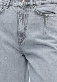 Vue de face d'un jean en denim gris clair montrant le bouton, la fermeture éclair, les passants de ceinture et les poches avant.
