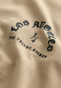 Beige stof med en glat tekstur har et sort grafisk print. Designet inkluderer "LOS ANGELES" og "THE VELVET EMBER" i forskellige skrifttyper.