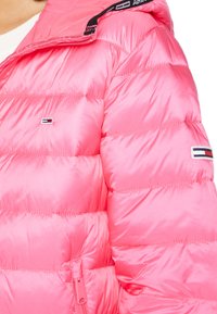 Jasnoróżowa puchowa kurtka o błyszczącej fakturze, z poziomymi wzorami pikowania, zapinana na zamek, z akcentami logo marki na rękawie i kołnierzu.