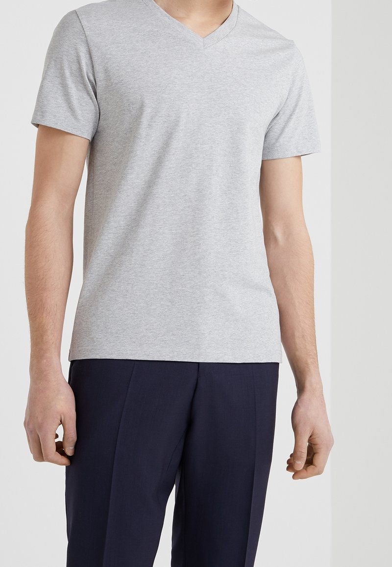 Homme portant un t-shirt uni gris clair à col en V et manches courtes ainsi qu’un pantalon bleu marine foncé, debout devant un fond clair.