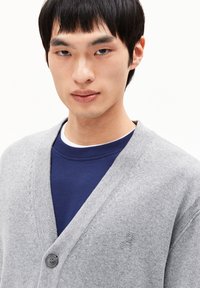 Gilet gris en tissu tricoté avec un col en V, fermeture à un bouton et un logo discret. Porté sur un t-shirt bleu à col rond.