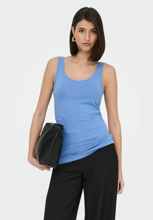 Woman with shoulder-length dark hair wearing blue sleeveless top and black pants, holding a black clutch bag by her side.