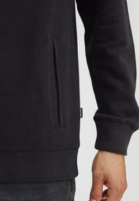 Black sweatshirt featuring a side seam pocket, ribbed cuffs, and hem. Soft texture with stitched detail and branded tag on the side.