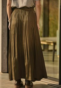 Jupe longue plissée vert olive avec une taille élastique, associée à des bottes marron foncé, présentée de dos dans un cadre intérieur moderne.