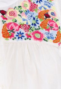 Bunte Blumen-, Schmetterlings-, Karotten-, Erdbeer- und Marienkäferstickerei auf weißem Stoff mit gerafftem Naht und Knöpfen.