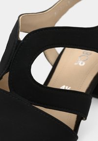 Μαύρο σανδάλι με υφή στην επιφάνεια, σχέδιο με ανοίγματα και δύο φαρδιές λουρίδες. Το εσωτερικό είναι λείο με ορατό λογότυπο. Χαμηλό τακούνι.