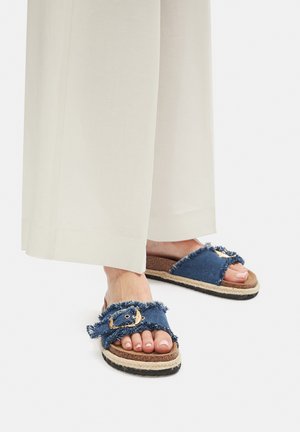 Sandalias tipo slide de mezclilla con bordes deshilachados, detalle de hebilla dorada y plataforma de yute. La suela es texturizada y oscura para mayor agarre.