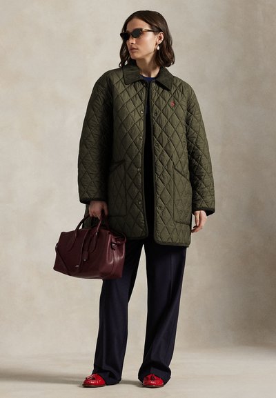 Naine kannab tumerohelist tepitud jopet, musti pükse, punaseid loafereid, päikeseprille, hoides suurt burgundia käekotti neutraalsel taustal.