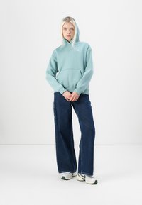 Lichtblauwe hoodie met een kangoeroezak en capuchon, gecombineerd met losse donkere denim jeans en witte sneakers met groene accenten.