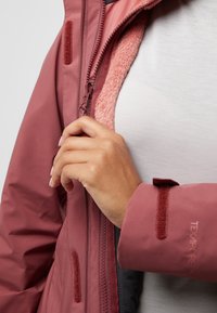 Veste imperméable couleur rose poussiéreux avec une douce doublure en polaire rose. Équipée d'une fermeture éclair et de velcro, avec des manches texturées.