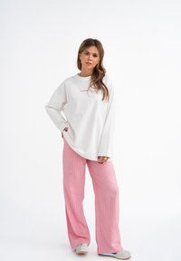 Oversized witte sweatshirt met roze tekst, gecombineerd met wijde roze gestreepte broek en grijze instapschoenen. Eenvoudig ontwerp met een casual pasvorm.
