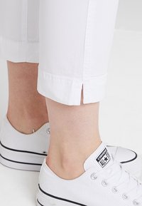 Pantalon blanc longueur cheville avec une petite fente latérale, porté avec des baskets blanches basses Converse All Star sur fond blanc uni.