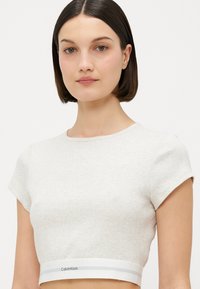 Kortärmad, croppad topp i ljusgrått ribbat tyg med en vit band i nederkanten som har en "Calvin Klein"-logga.