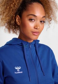 Blauwe ritsup hoodie van zacht materiaal, met een capuchon met trekkoord en een wit geborduurd "hummel" logo op de borst.