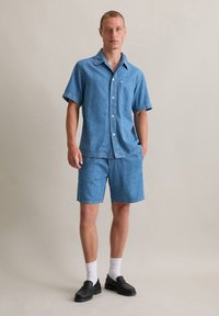 Lichtblauwe denim kortekool shirt met een zak, gecombineerd met bijpassende denim shorts. Het model draagt witte sokken en zwarte instappers.