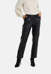 Pantalon en similicuir noir avec une taille élastique et un cordon de serrage. Coupe fuselée, texture brillante, associé à des bottes noires à talons hauts.