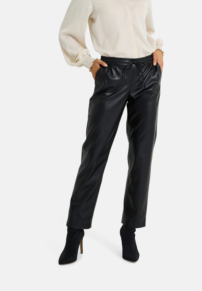 Pantalon en similicuir noir avec une taille élastique et un cordon de serrage. Coupe fuselée, texture brillante, associé à des bottes noires à talons hauts.