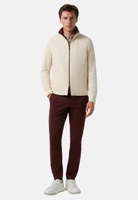 Beige Zip-up-Jacke mit maroonfarbenem Kragen, kombiniert mit burgunderfarbenen Hosen und weißen Sneakern. Die Jacke hat seitliche Taschen und eine glatte Textur.