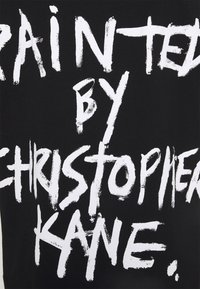 Texte blanc manuscrit sur fond noir indiquant « "Painted by Christopher Kane" » en style de coup de pinceau gras et irrégulier.