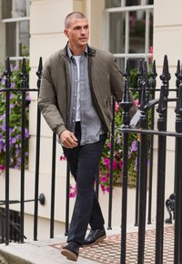 Olivgrüne Bomberjacke über einem grauen karierten Hemd, kombiniert mit schwarzen Hosen und dunklen Sneakern, auf einem Weg mit Blumen im Hintergrund gehend.