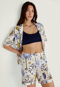 Woman with long dark hair wearing a navy crop top and matching patterned open shirt and shorts with celestial and botanical designs.